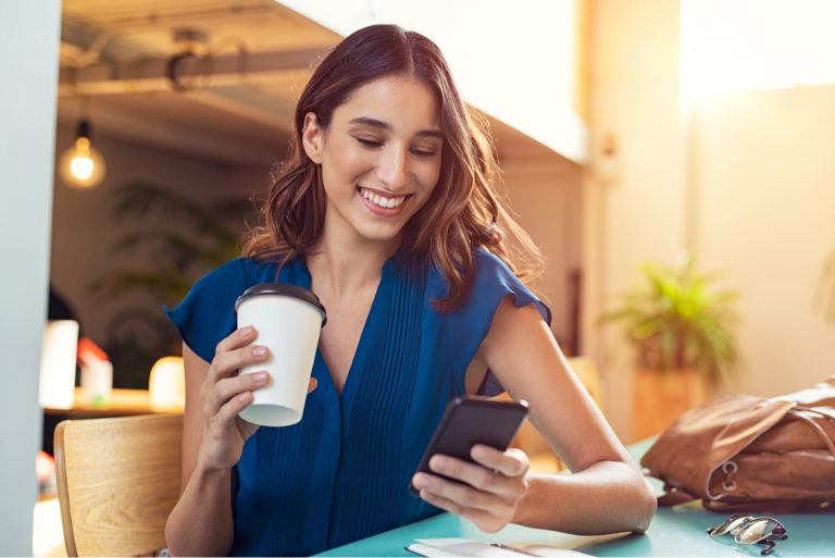 eine lächelnde Frau, die an einem Tisch sitzt und Kaffee trinkt und ein Telefon drückt