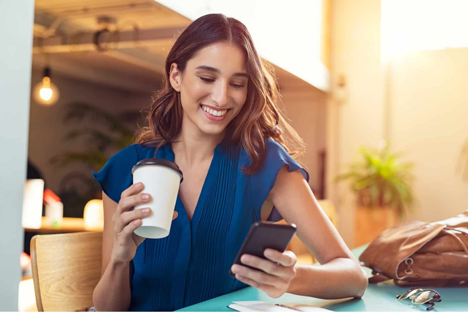 eine lächelnde Frau, die an einem Tisch sitzt und Kaffee trinkt und ein Telefon drückt