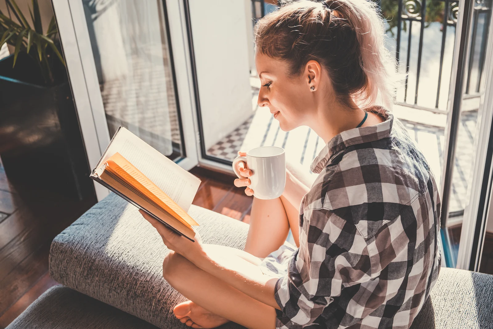 Frau sitzt und trinkt Tee, während sie ein Buch am offenen Fenster liest