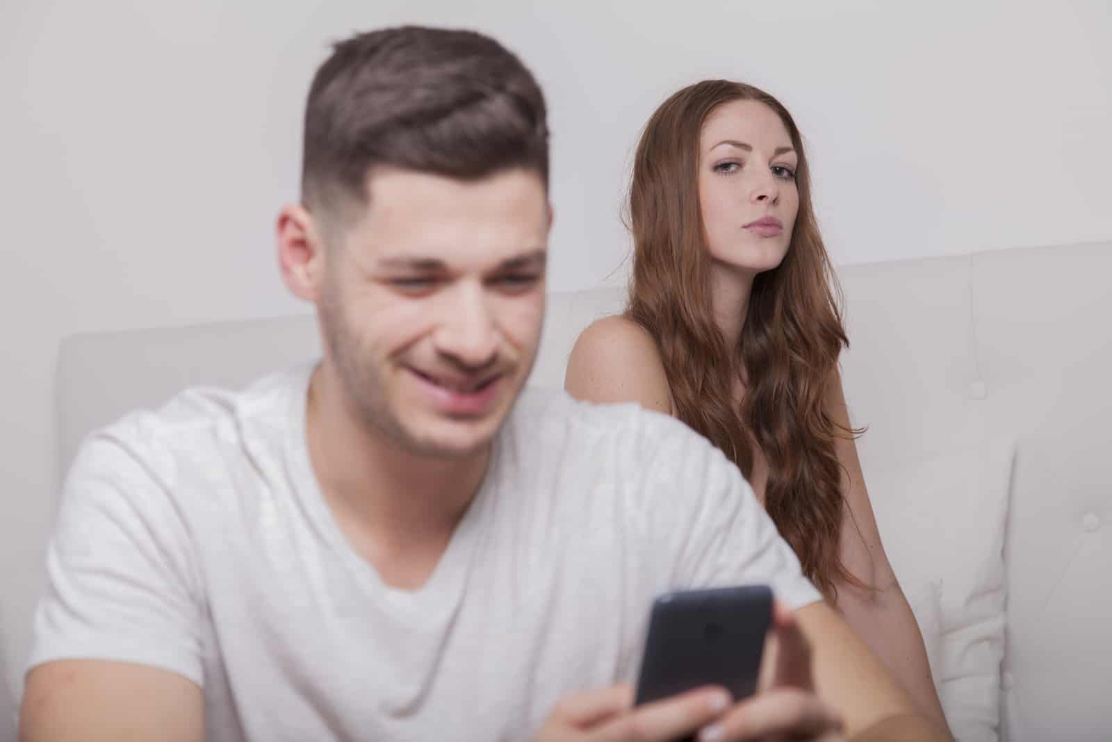 Der Mann erhält eine Nachricht auf dem Smartphone, während die Frau eifersüchtig aussieht
